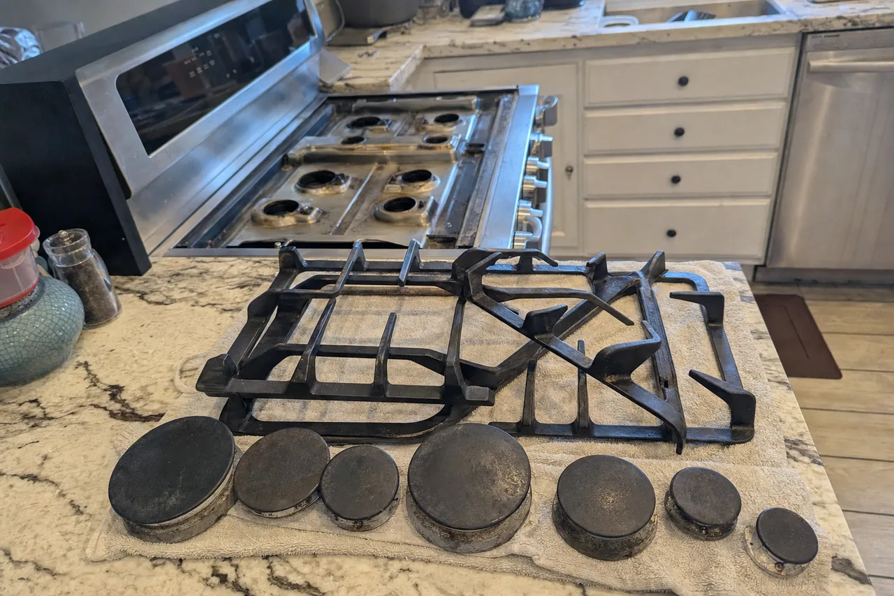 Cast-iron grates and a row of burner caps laid out on a granite island in front of a stainless rangetop with its burner bases exposed