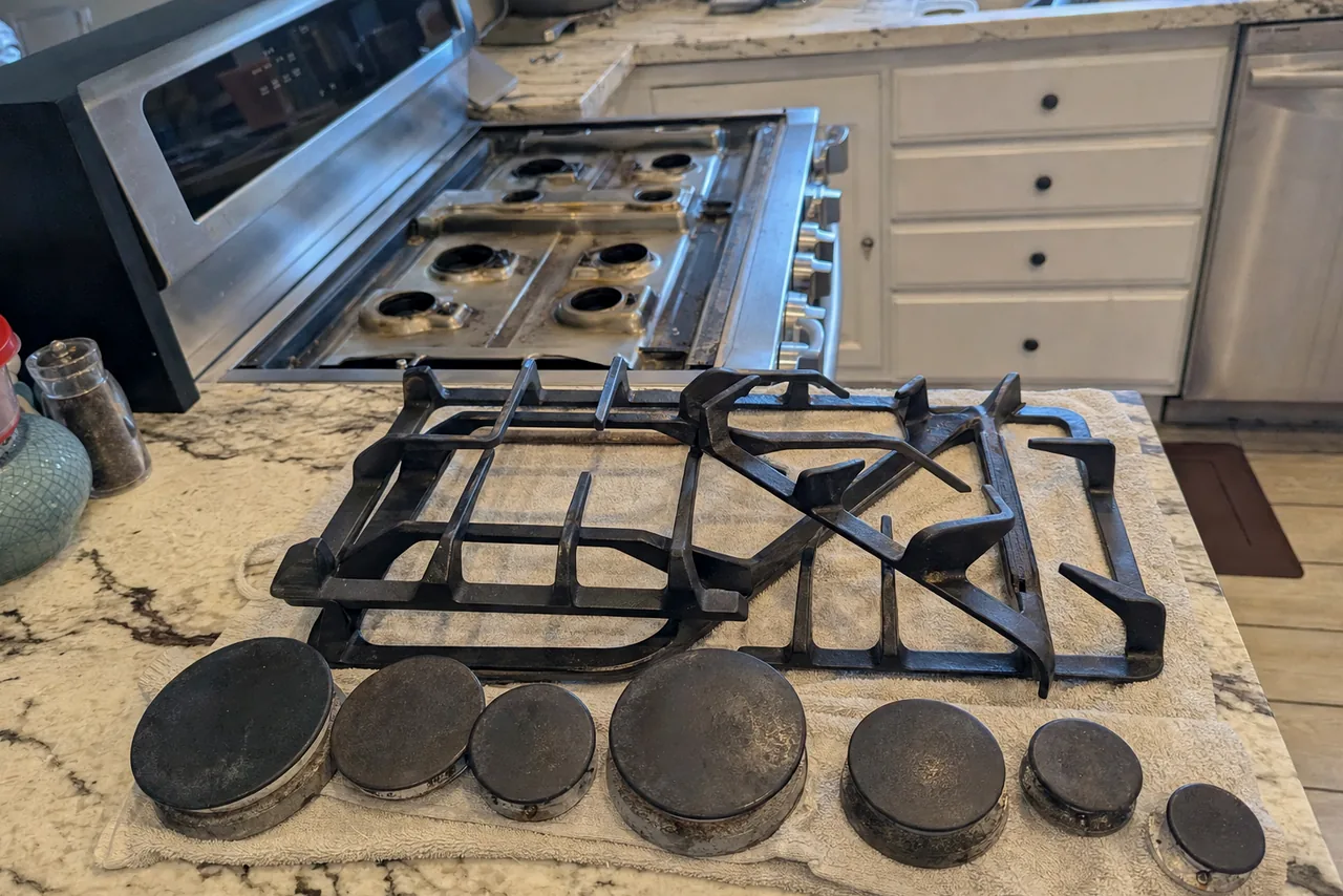 A stainless rangetop stripped down to its burner bases, the grates and caps stacked on the counter beside it